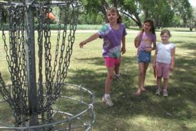 3 young girls near disc golf basket and one of them is throwing a disc at the basket